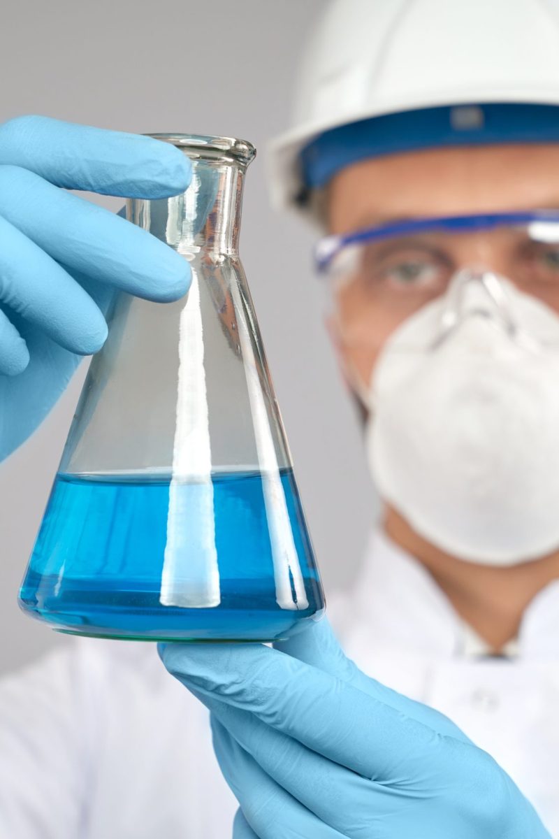 Close up of flask with blue liquid. Close up of young chemist in glasses, helmet and mask doing experiment in laboratory, isolated on grey. Selective focus of flask with blue liquid in hand of scientist in gloves and white lab coat.
